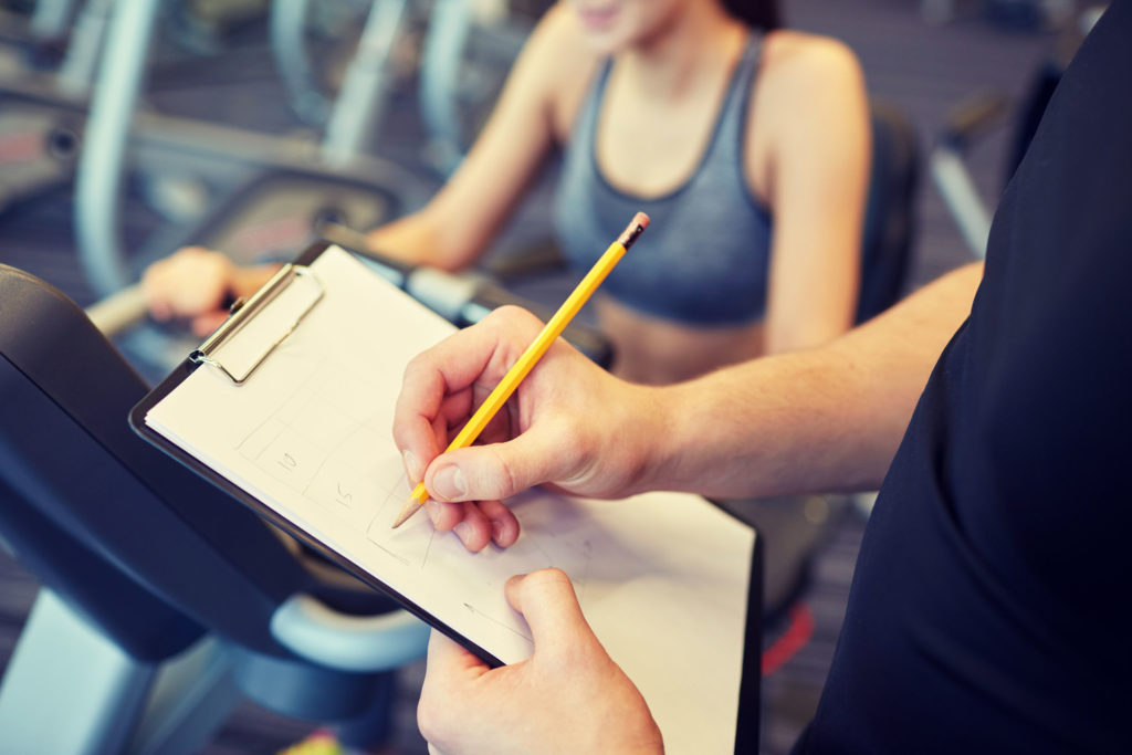 49090294 - sport, fitness, lifestyle, technology and people concept - close up of trainer hands with clipboard writing and woman working out on exercise bike in gym